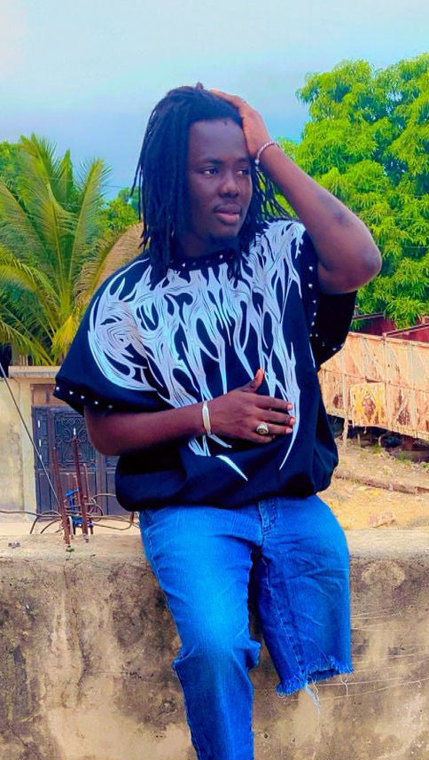 Amzee Prince du Sud wearing a black and white patterned shirt with blue jeans sitting on a ledge outdoors.